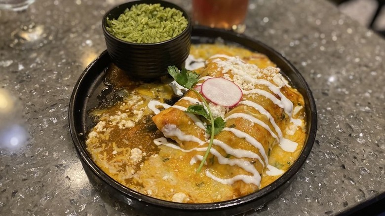 A round plate with two cheesy enchiladas in it, drizzled with sour cream and garnished with a radish slice and cilantro sprig with green rice on the side