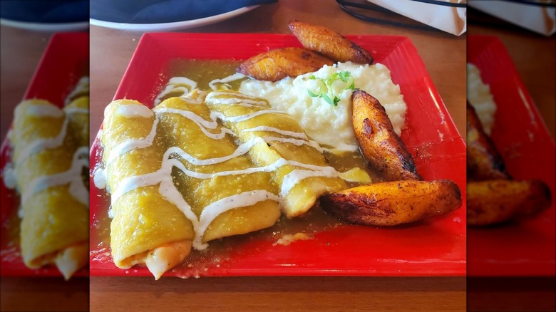 Three enchiladas on a square red plate smothered in verde sauce and drizzled with crema, with cheesy rice and fried plantains on the side