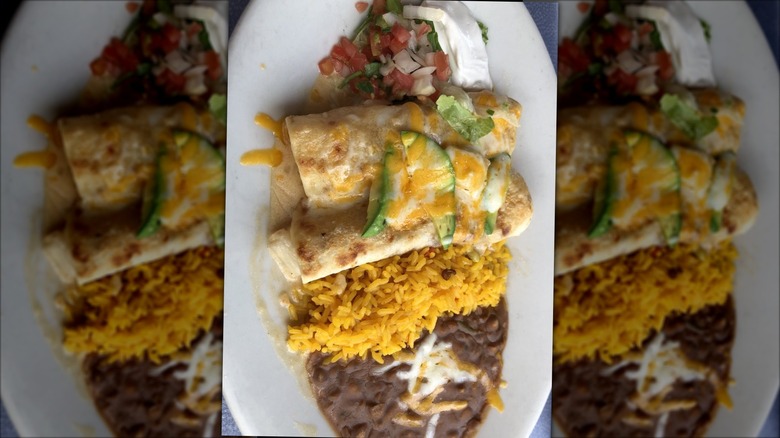 An overhead shot of two enchiladas on a white plate with melted cheese and avocado on top; pico, sour cream, rice and beans on the side