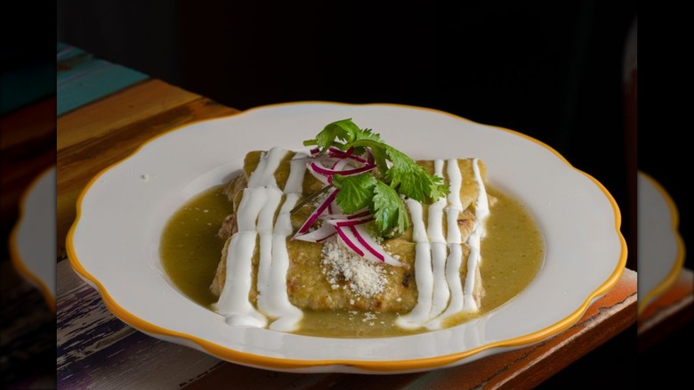 Three enchiladas in a white dish covered in green sauce and drizzled wtih sour cream and Cotija cheese, garnished with sliced radishes and cilantro