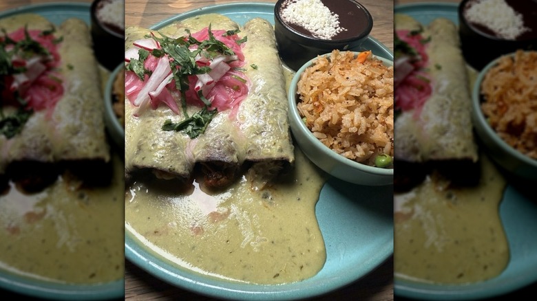 Three enchiladas on a light blue plate covered in Suiza sauce with Mexican rice and black beans on the side