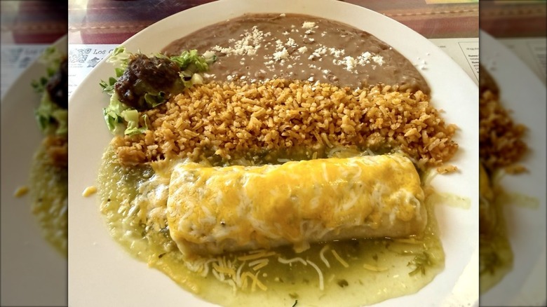 A single enchilada on a white plate covered in cheese and green sauce with Mexican rice and refried beans on the side