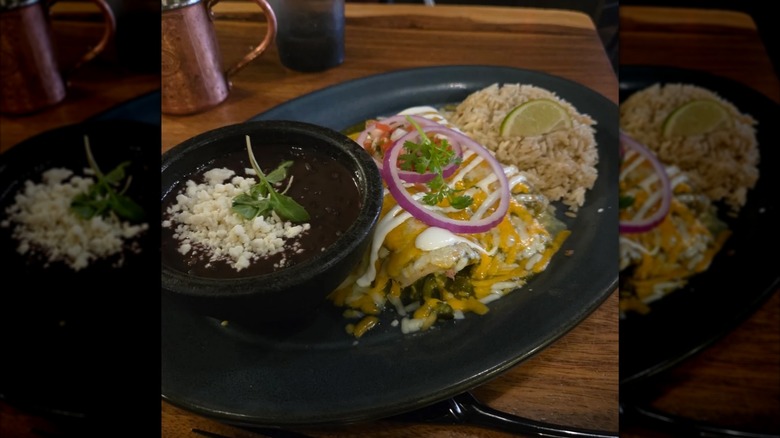 Two enchiladas on a black plate covered in cheese and sour cream, garnished with cilantro, pico, and sliced red onion, served with black beans and rice on the side