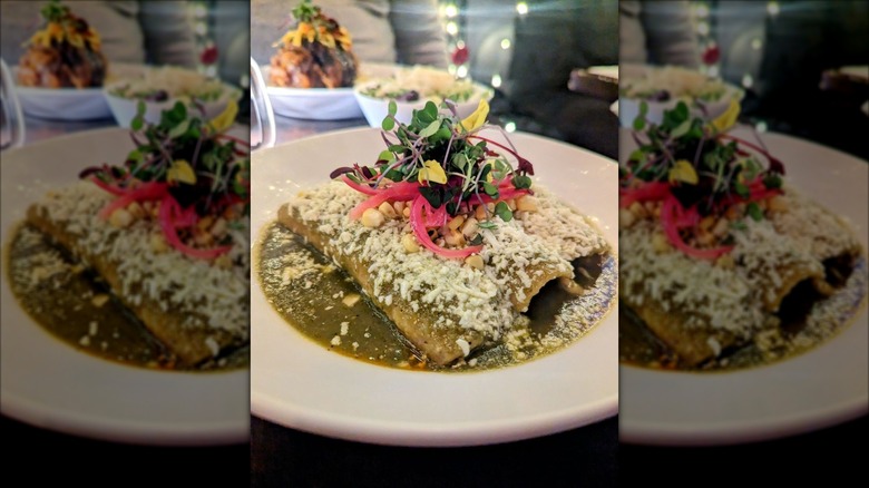 Three enchiladas on a white plate smothered in a green sauce, topped with queso fresco, and garnished with corn niblets, red onions, and micro greens
