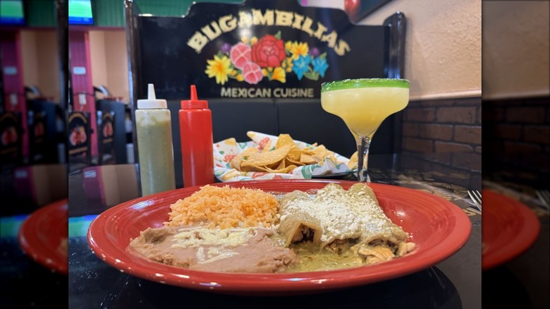 A red plate on a table with three enchiladas and sides, a margarita, chips, and the restaurant logo are in the background
