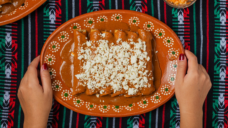 Six enchiladas smothered in red sauce and queso fresco on a terra-cotta-colored platter