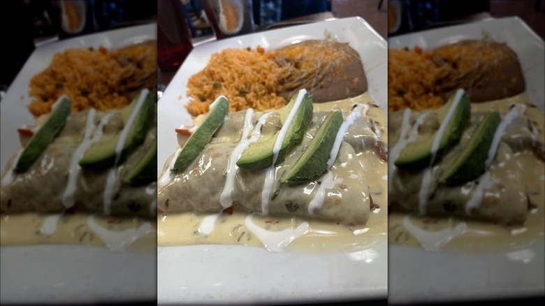 Two enchiladas on a rectangular white plate covered in a cream sauce, drizzled with sour cream, and topped with sliced avocados; rice and refried beans on the side
