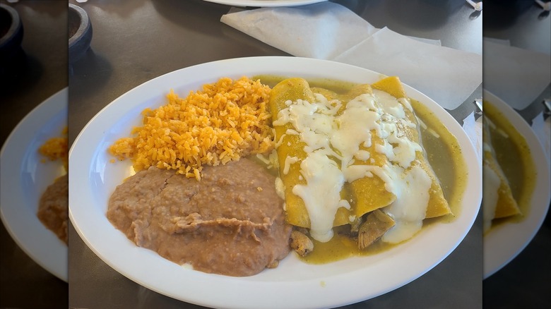 Three enchiladas on a white plate covered in verde sauce and melted cheese with rice and refried beans on the side
