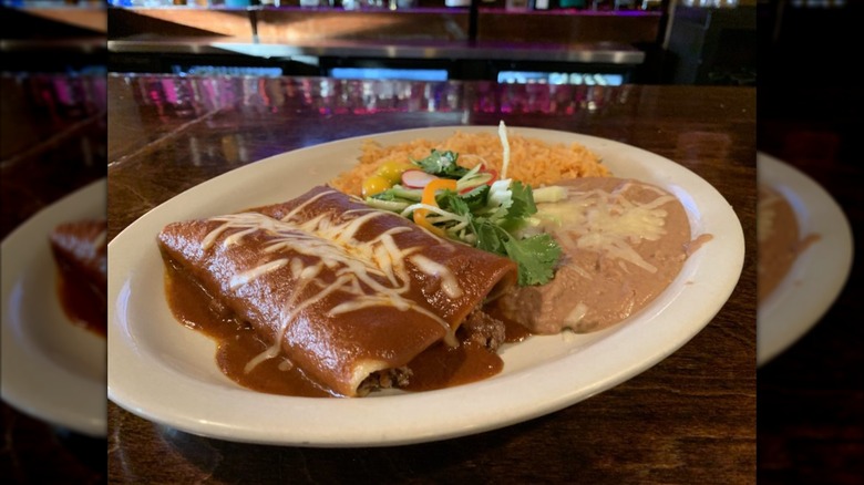 Two meat-filled enchiladas on a white plate covered in red sauce and melted cheese, garnished with cilantro and sliced radish; rice and refried beans on the side