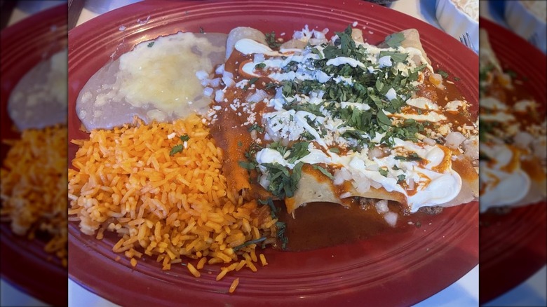 Four enchiladas covered in red sauce, cilantro, queso fresco, and sour cream on a red plate with Mexican rice and refried beans with cheese on the side