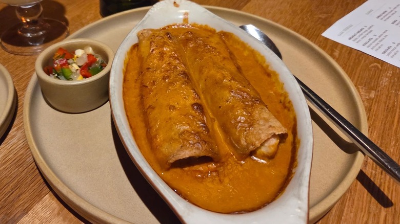 Two enchiladas in a white boat-style dish covered in a creamy orange sauce; a ramekin of fresh salsa is on the side