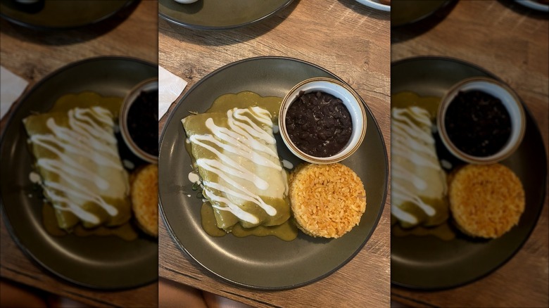 Three enchiladas covered in green sauce, melted cheese, and sour cream on a dark plate with black beans and rice on the side