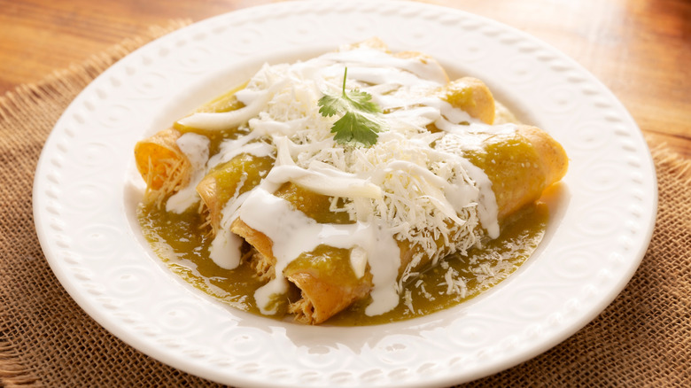 Four chicken enchiladas smothered in green sauce, sour cream and white cheese garnished with a spring of cilantro on a white plate