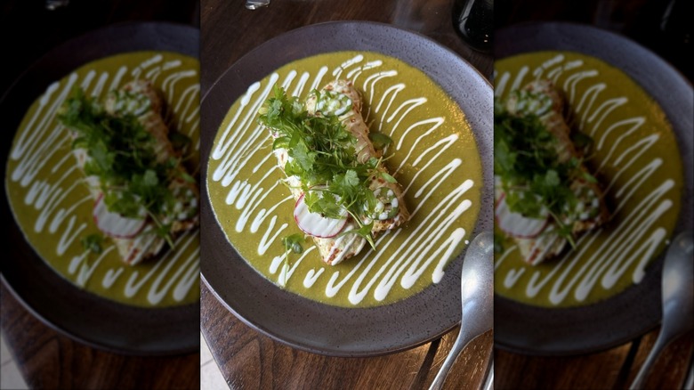 Two enchiladas on a dark plate smothered in verde sauce, drizzled with sour cream and topped with sliced radishes and cilantro