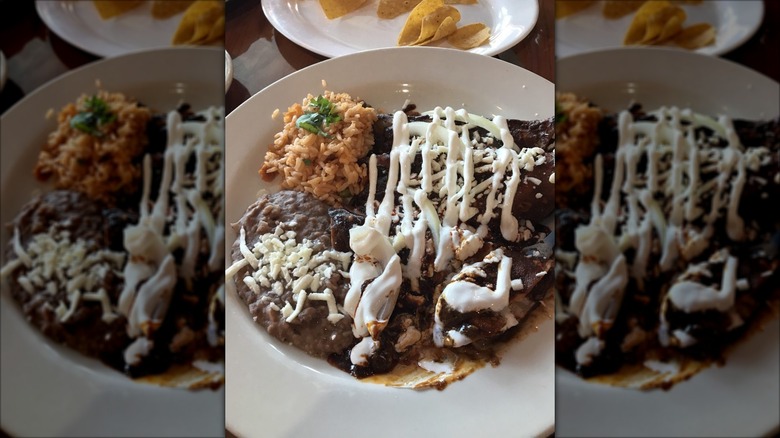 Partially eaten enchiladas smothered in mole sauce with white cheese and a drizzling of sour cream; refried beans and rice on the side