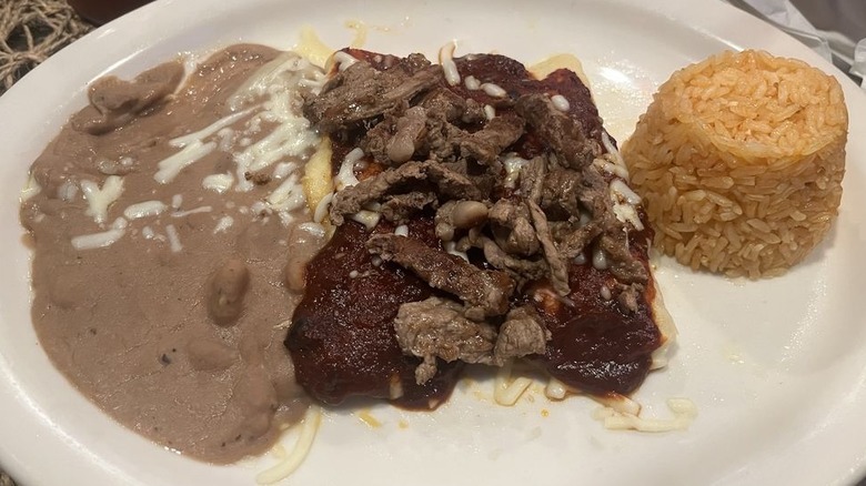 Two enchiladas on a white plate covered in red sauce and chopped steak with refried beans and Mexican rice on the side