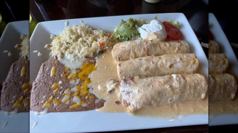 Three enchiladas on a white rectangular plate covered in a cheese sauce with refried beans, rice, guacamole, sour cream and a tomato slice on the side