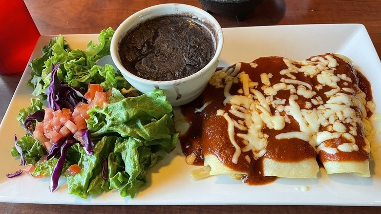 Three enchiladas covered in red sauce and melted cheese on the right side of a white rectangular plate with a side salad and black beans on the side