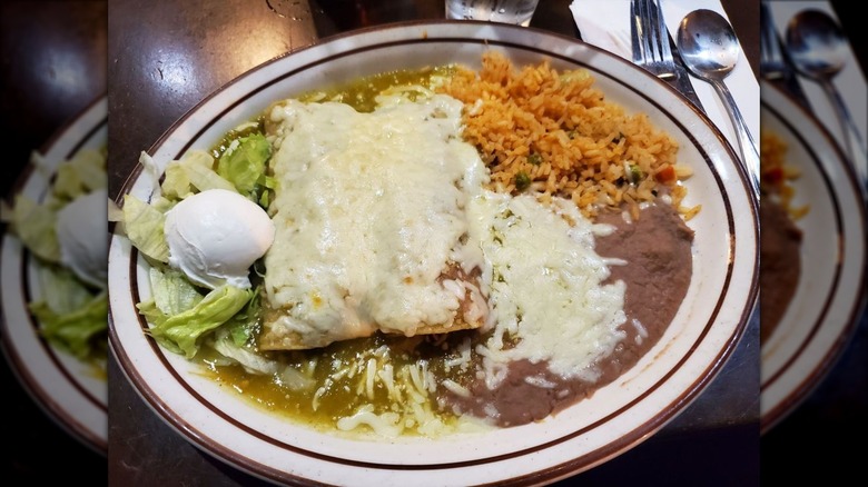 Two enchiladas covered in verde sauce and melted cheese on a round plate with lettuce, sour cream, Mexican rice, and refried beans on the side