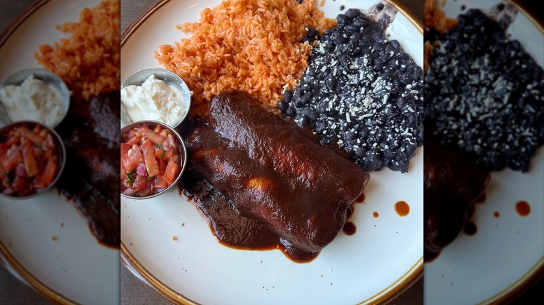 Two enchiladas covered in a rich red sauce with pico, sour cream, Mexican rice, and black beans on the side