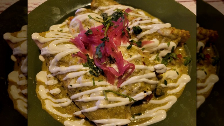 Three enchiladas on a dark green plate covered with verde sauce, drizzled with sour cream, and garnished with pickled red onion and cilantro