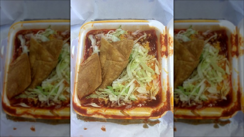 Enchiladas in a styrofoam to-go container, barely visible under a tortilla chips, a pile of lettuce, and red sauce