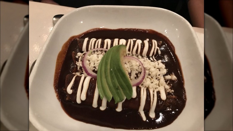 A dish of three enchiladas smothered in mole sauce, drizzled with sour cream, sprinkled with queso fresco, and garnished with sliced red onion and avocado