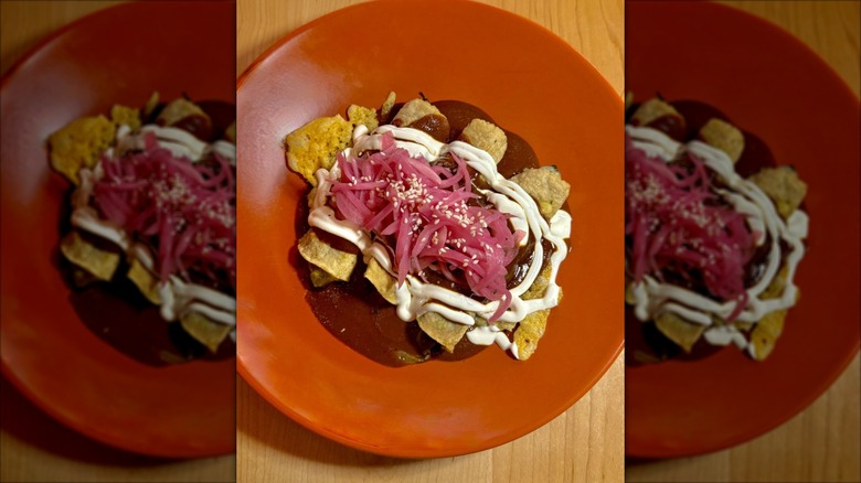Three enchiladas on an orange plate covered in dark red sauce, drizzled with sour cream, and garnished with pickled red onion and queso fresca