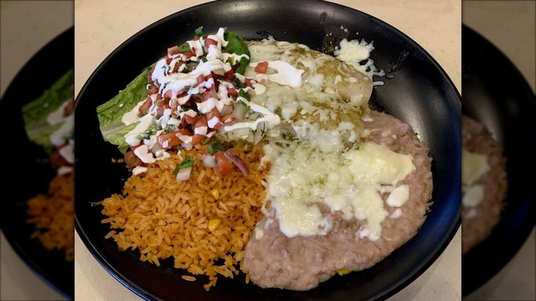 Two enchiladas verde on a black plate with a small salad, Mexican rice, and refried beans; drizzled with sour dream and melted cheese