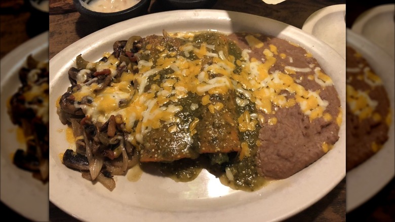Two enchiladas covered in green sauce and cheese on a white plate with grilled veggies and cheesy refried beans on the side