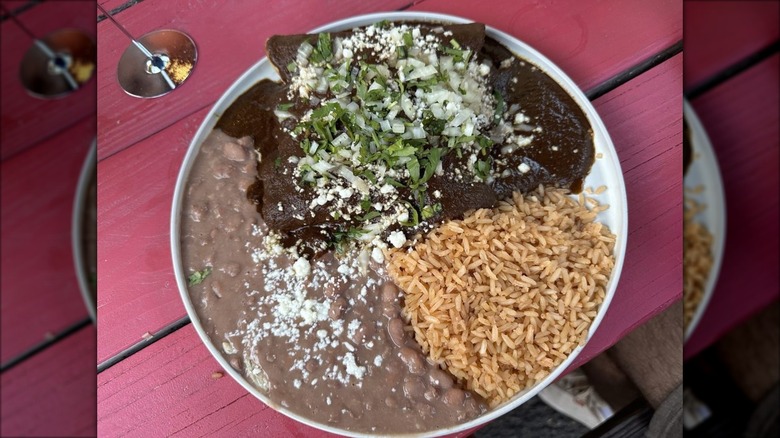 Enchiladas mole garnished with queso fresco, chopped cilantro and onions on a white rimmed plate with Mexican rice and refried beans on the side