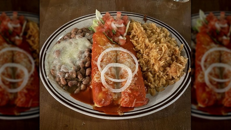 Two enchiladas with red sauce, garnished with sliced onion and pico on a white plate with two black rings around the rim; cheesy pinto beans and Mexican rice on the side