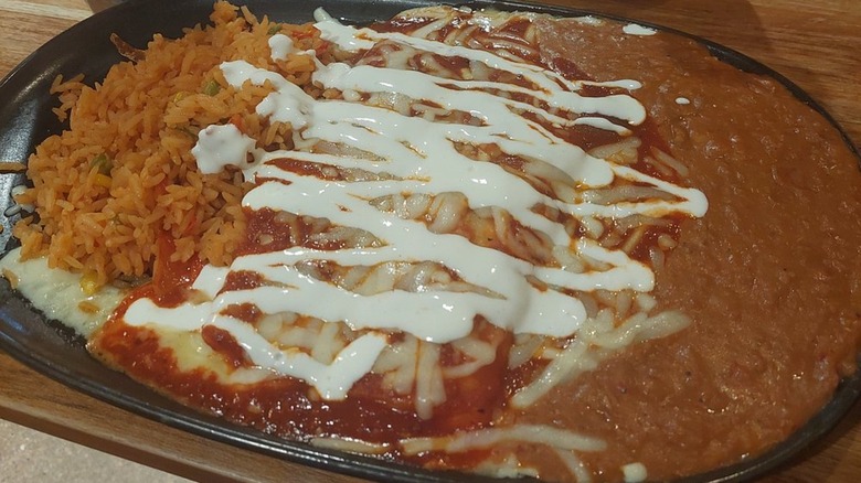 Two enchiladas on a black plate covered in red sauce, melted cheese, and drizzles of sour cream with Mexican rice and refried beans on the side