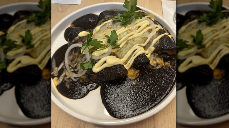 Three enchiladas on a rimmed white plate covered in black mole sauce with melted cheese and drizzles of yellow sauce; garnished with sliced white onions and cilantro