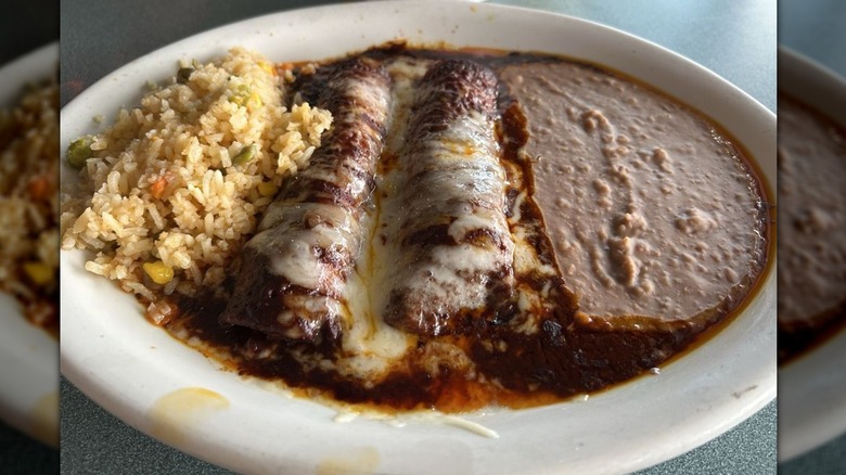 Two enchiladas mole covered in melted cheese on a white plate with Mexican rice and refried beans on the side