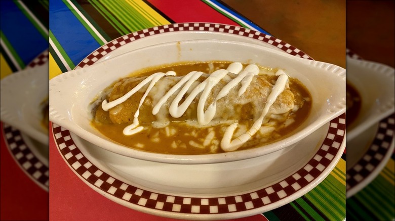 An enchilada in a white ceramic food boat, covered in orange sauce with melted cheese and sour cream drizzle; vibrant tablecloth in background