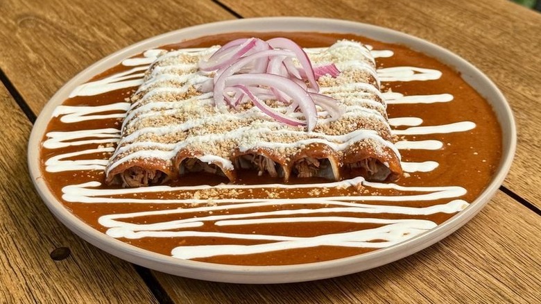 Five enchiladas on a white rimmed plate covered in orange sauce, sprinkled with queso fresca, drizzled with sour cream, and garnished with red onion