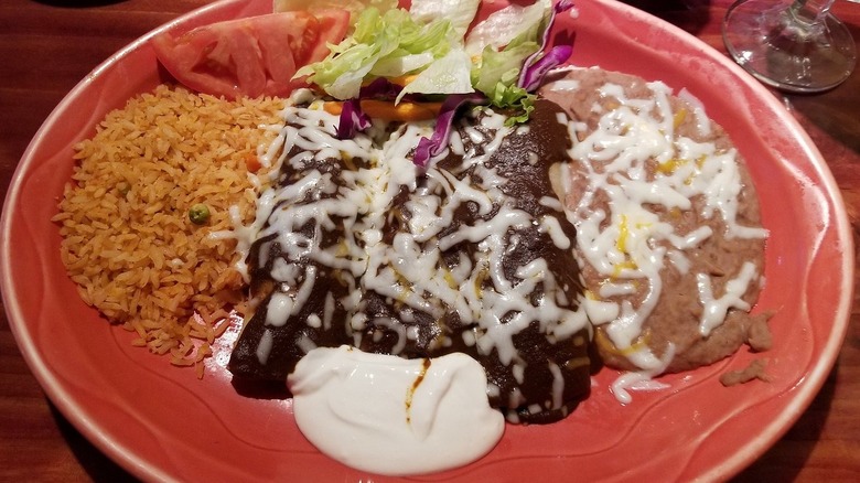 Three enchiladas on a red plate covered in mole sauce and melted cheese; Mexican rice, a small salad, refried beans, and sour cream on the side