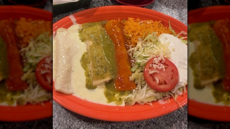 Three enchiladas on a red plate; one covered in cheese sauce, one in green sauce, and one is red sauce; Mexican rice, shredded lettuce, sour cream, and a tomato slice on the side