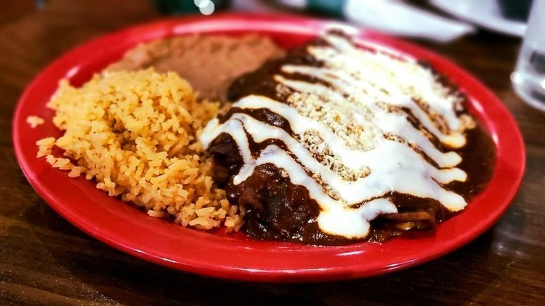Enchiladas mole on a red plate sprinkled with queso fresca and drizzled with sour cream; Mexican rice and refried beans on the side