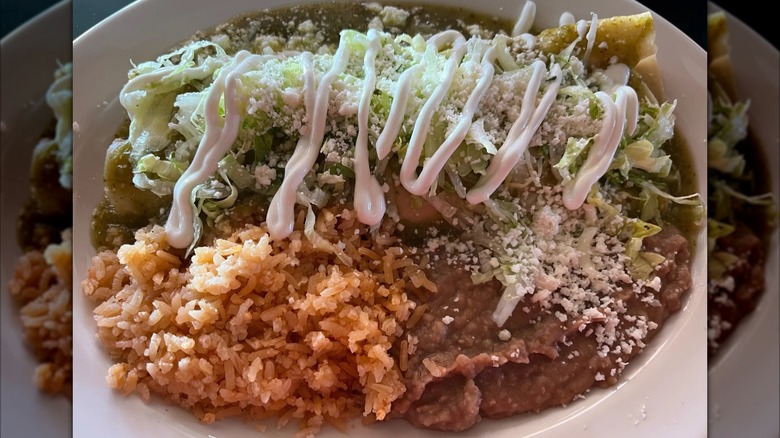 An enchilada on a white plate covered in green sauce, lettuce, and queso fresca drizzled with sour cream; Mexican rice and refried beans on the side