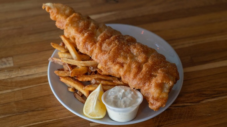 plate with large piece of fried fish over fries served with lemon wedge and plastic ramekin of tartar sauce