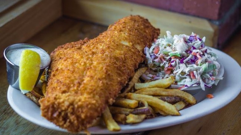 large piece of fried fish with fries next to cole slaw and tartar sauce with lemon wedge