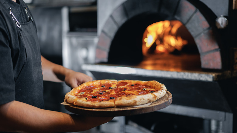 A man holding a pepperoni pizza fresh out of the wood-fired oven in the background