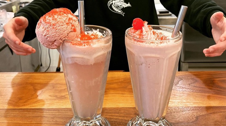 Two pink milkshakes in glasses with metal straws, extra scoop on one, cherry and whipped cream on other.