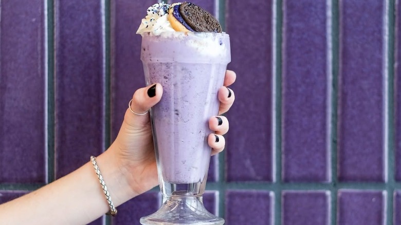 Purple milkshake in a glass, topped with an Oreo cookie, against purple wall.