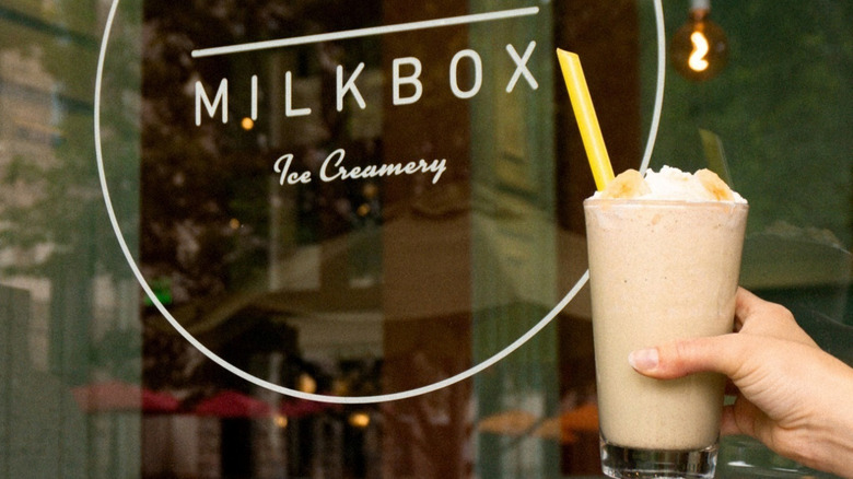 Milkshake topped with banana slices and whipped cream with yellow straw, in front of Milkbox window.
