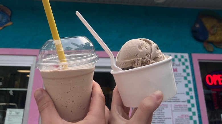 Two hands holding up a milkshake and an ice cream cup in front of DeNucci's.