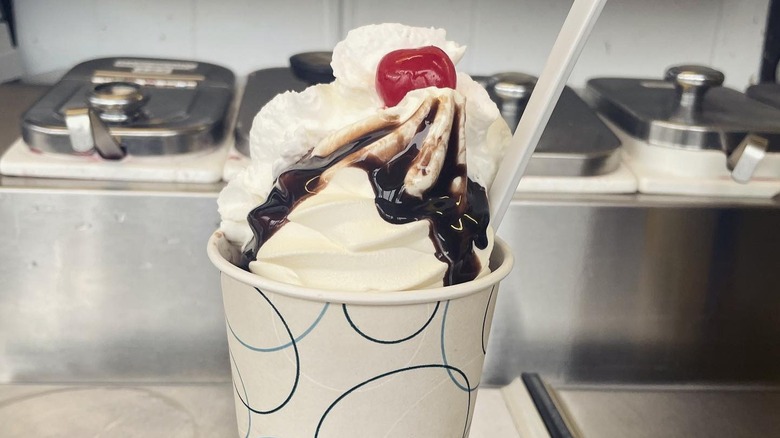 Milkshake topped with whipped cream, chocolate syrup and cherry in paper cup, in kitchen.