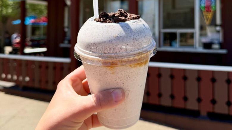 Hand holding full milkshake in plastic cup with cookie topping.
